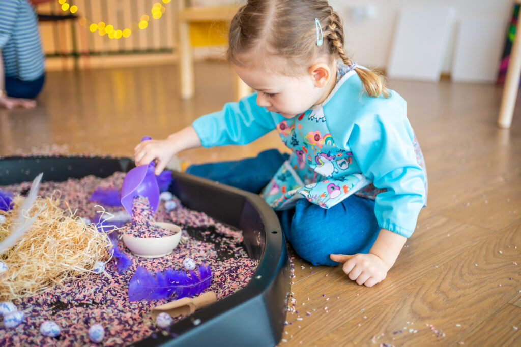 Niña participando en una actividad sensorial, explorando diferentes texturas y materiales como arroz coloreado en un espacio educativo.